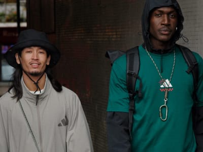 Two individuals, one wearing a gray hooded jacket and the other wearing a green shirt, stand in an urban setting with buildings and structures visible in the background.