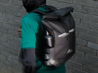 A black and gray backpack with reflective straps and pockets is shown against a brick wall background.