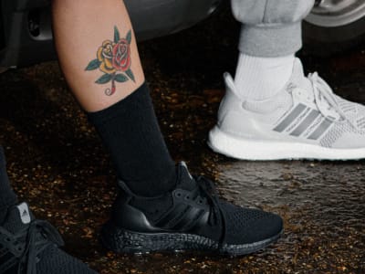 The image shows a person's arm with a rose tattoo, wearing black boots, and standing on a dirt or gravel surface.