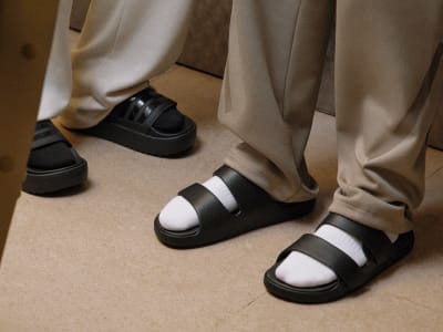 The image shows a pair of black and white sandals on a wooden floor, with beige curtains visible in the background.
