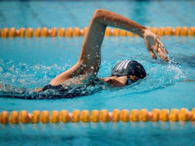 A swimmer in a black swimsuit is gliding through the water in a swimming pool, with a bright blue background and yellow lane markers visible.