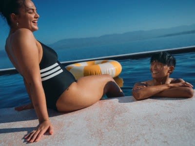 The image shows a woman in a black and white striped swimsuit reclining on a boat, with a man in the background also reclining on the boat, against a backdrop of a serene blue ocean and sky.