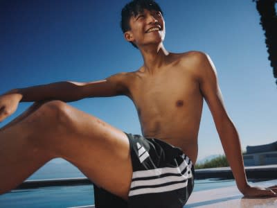 A shirtless young man sits on the edge of a pool, smiling against a clear blue sky.