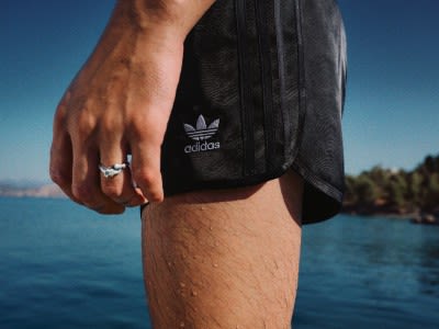 The image shows a person's lower body wearing black Adidas shorts standing near a body of water with a clear blue sky in the background.
