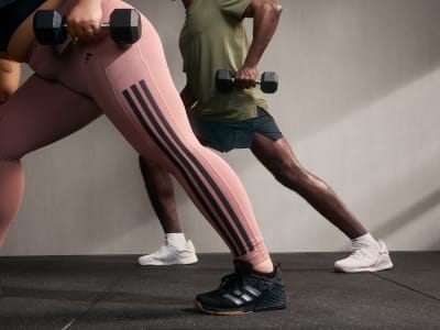 The image shows a person wearing pink and white athletic leggings and holding dumbbells, standing in a dimly lit room.