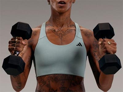A person wearing a white tank top and holding two black dumbbells, standing against a plain background.