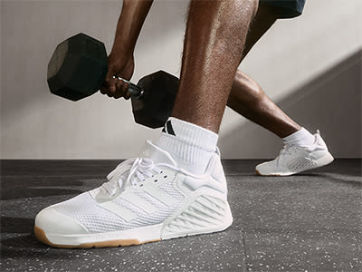 The image shows a person's lower body and feet, wearing white athletic shoes, lifting weights in a gym setting.