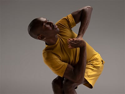 A person in a yellow shirt is captured in a dynamic pose, with their arms raised and body twisted, against a neutral gray background.