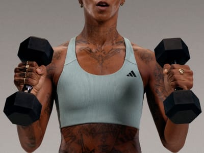 A muscular person wearing a white tank top and holding two black dumbbells, standing against a plain gray background.