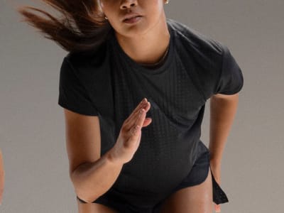 A person wearing a black top with their hair blowing in the wind, standing against a plain gray background.