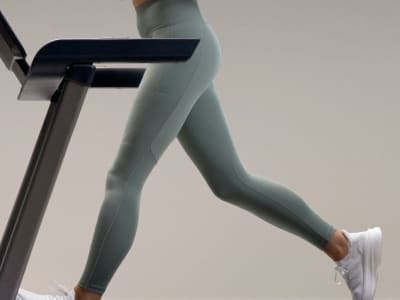 The image shows a person's lower body wearing light-colored athletic leggings and white sneakers, standing on what appears to be a piece of exercise equipment in a minimalist, neutral-toned setting.
