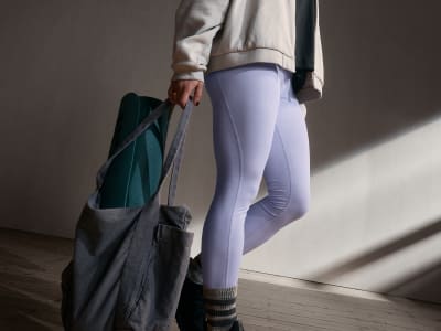 A person wearing light-colored pants and a gray jacket, holding a dark-colored bag, stands in a dimly lit room with a wooden floor.