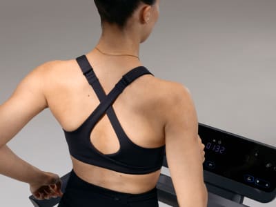 The image shows the back of a person wearing a black sports bra with a criss-cross design, standing in front of a plain gray background.