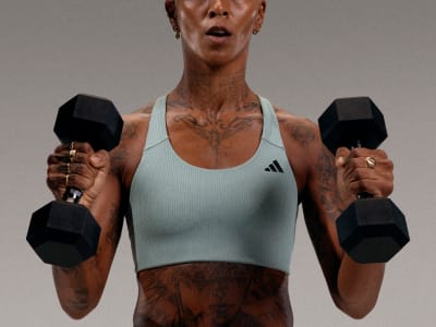 A muscular individual wearing a light-colored athletic top and holding two black dumbbells, standing against a plain background.