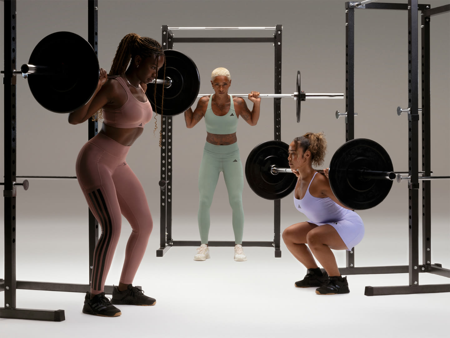 The image depicts three women in athletic attire engaged in weight training exercises in a gym setting, with various weightlifting equipment and racks visible in the background.