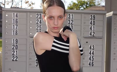 A young woman with blonde hair stands in front of a row of lockers, wearing a black and white striped top and a serious expression on her face.