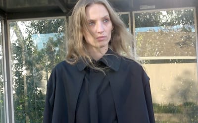 A young woman with long blonde hair stands in front of a glass window, wearing a dark-colored coat and looking directly at the camera.