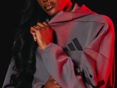 A person wearing a bright red hooded jacket stands in a dark background, with the Adidas logo prominently displayed on the jacket.
