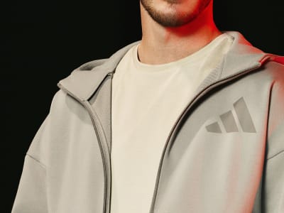 A close-up view of a person wearing a light-colored hooded jacket with the Adidas logo visible on the front, set against a dark background.