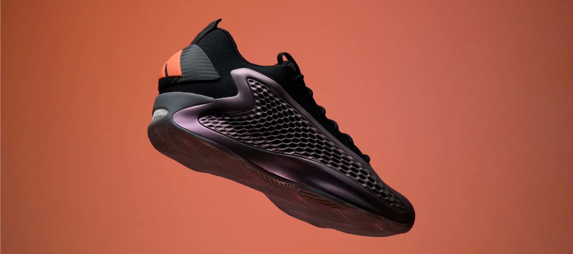 A sleek, futuristic-looking black and gray sneaker with a textured, angular design stands against a vibrant orange background.