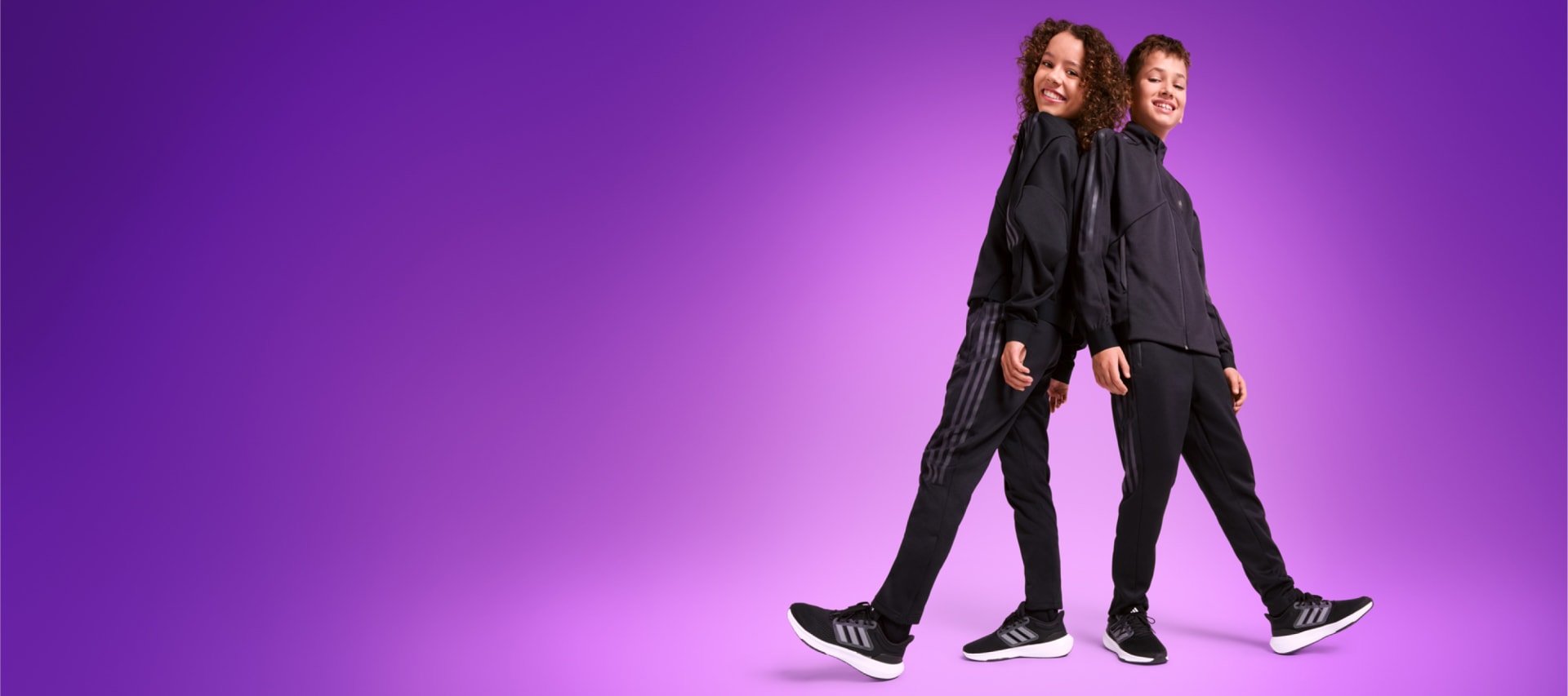 Two children, one male one female, stand smiling back to back in black adidas tracksuits and sneakers.