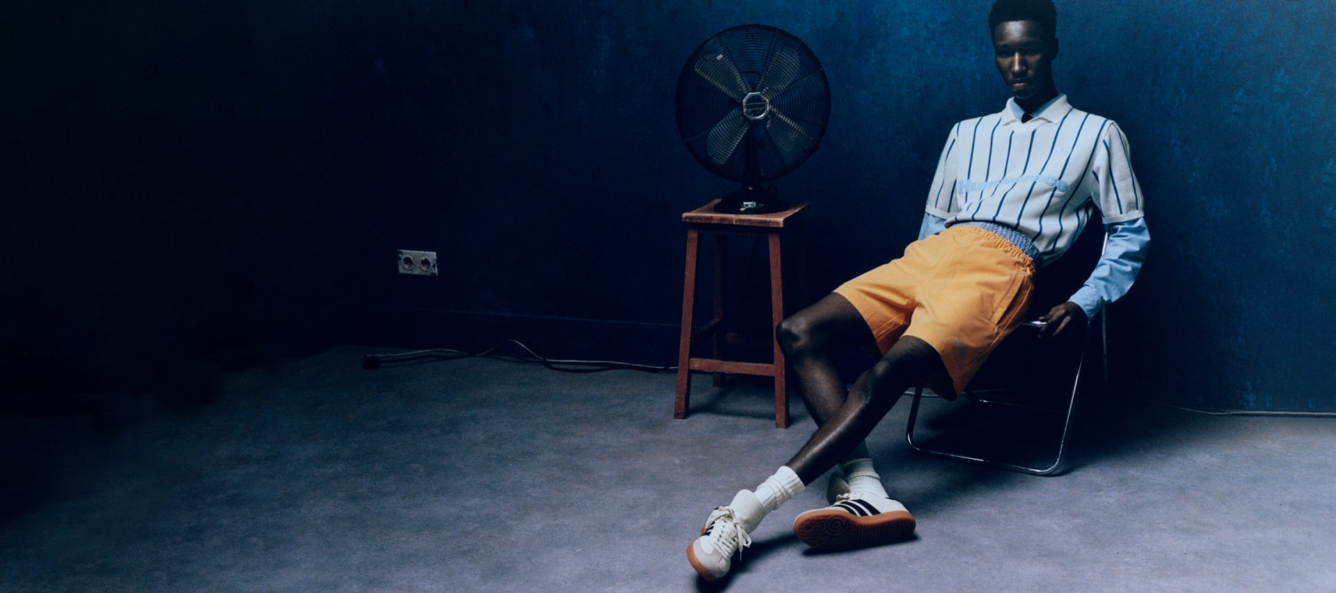 Model wearing blue shirt and orange shorts reclines on chair in dark room