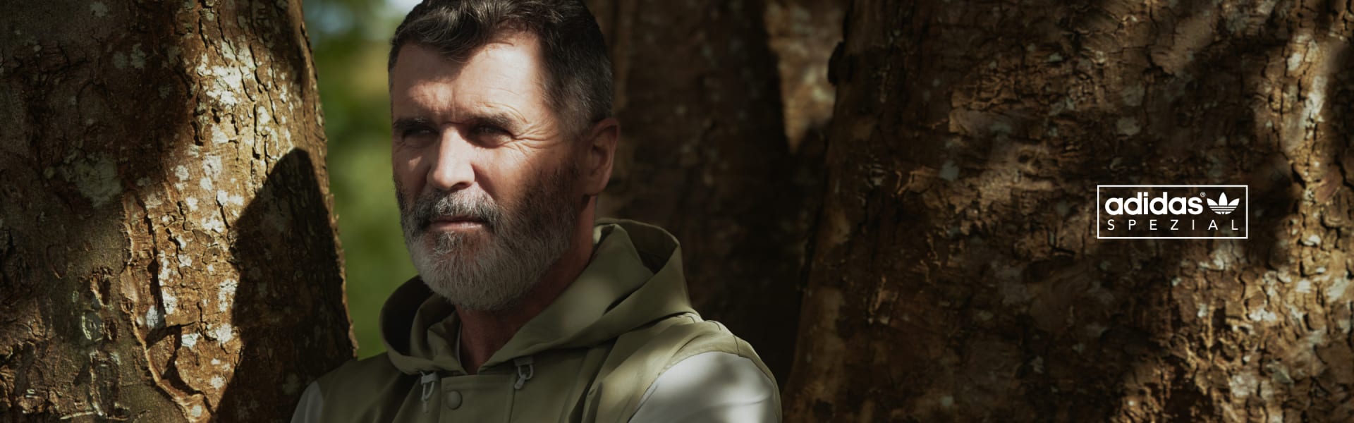 Roy Keane leans on a tree with crossed arms and a slight smile, hiding from the sun in a green and gray Spezial jacket.