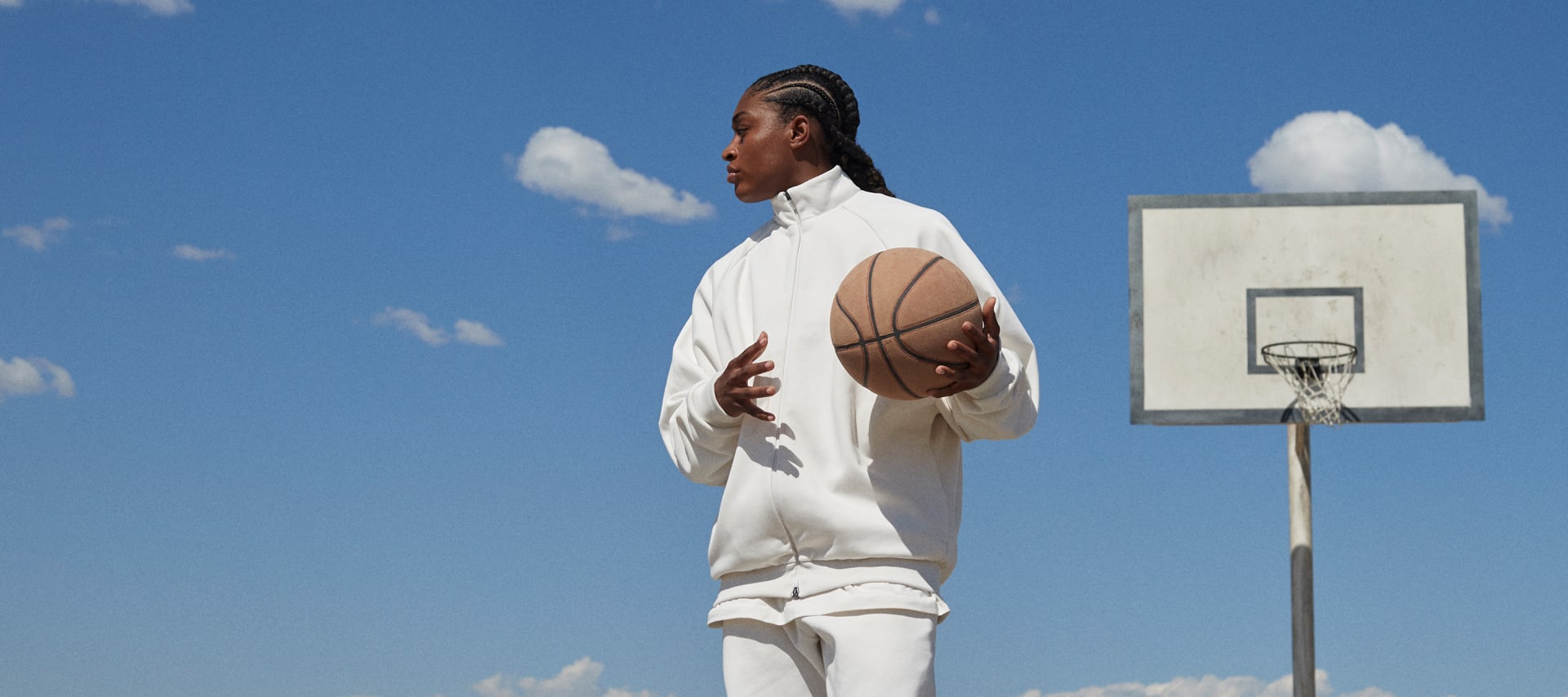 A woman looking to her right, wearing a white jacket and sweatpants and holding a basketball in her left hand, standing in front of a basketball goal and bright blue sky.