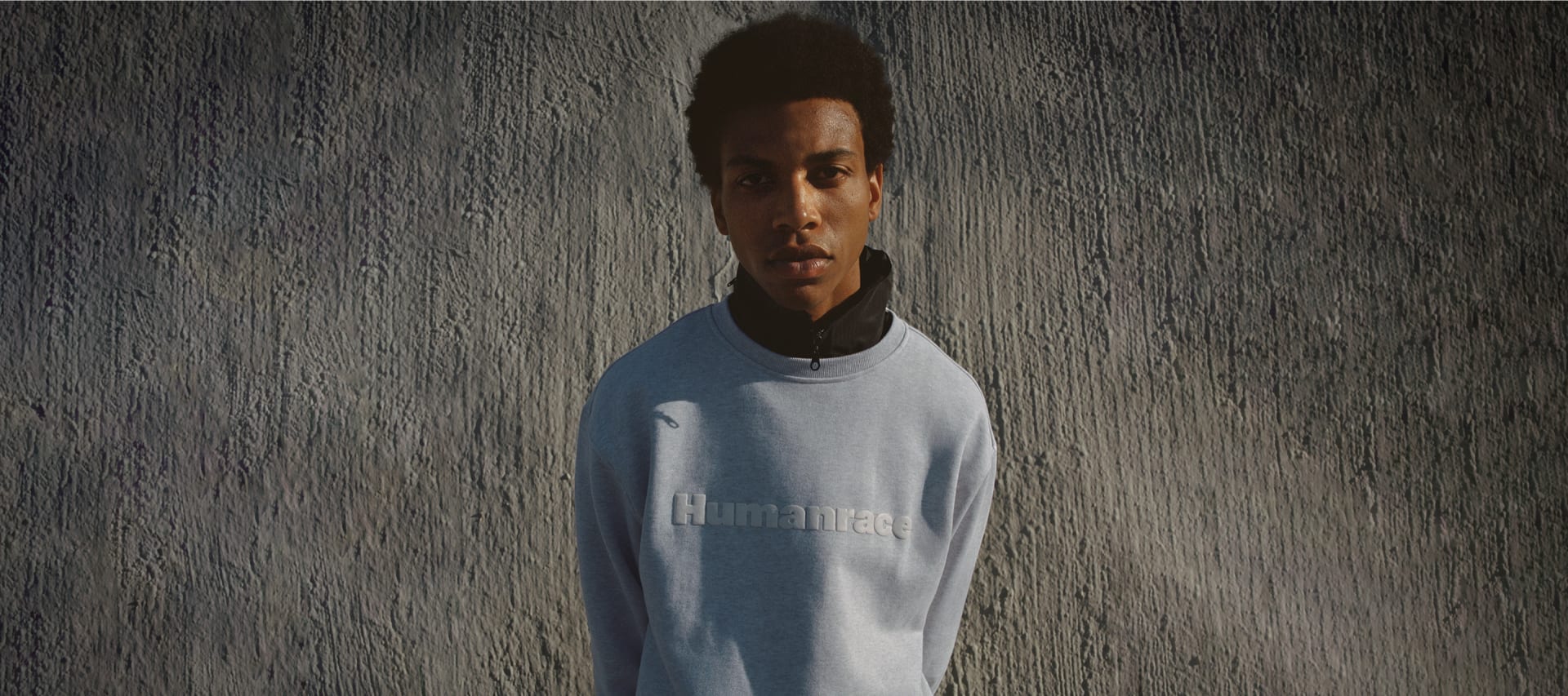 Model looks to camera wearing gray jumper against concrete background.
