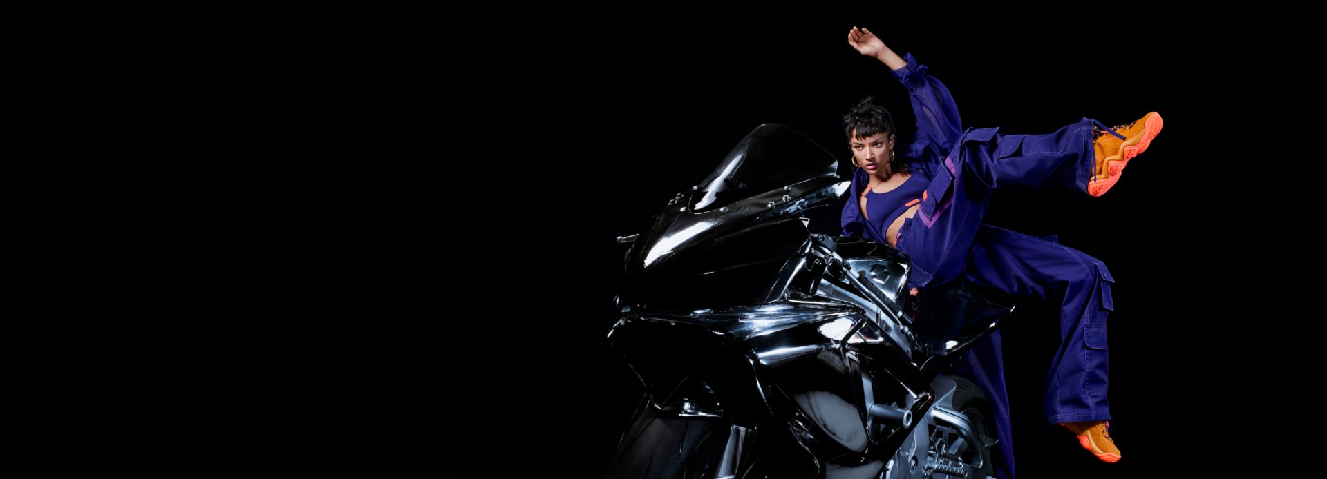 A female model is photographed posing on a motorbike in the adidas x IVY PARK collection against a black backdrop.