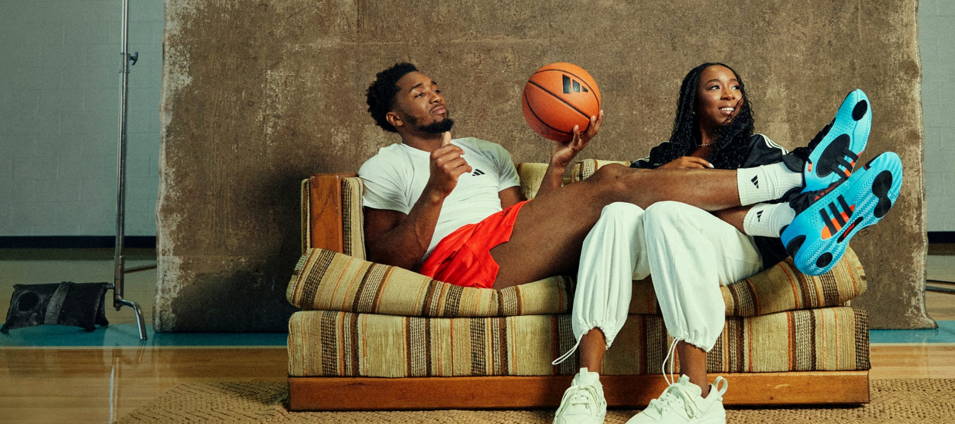 Donovan Mitchell relaxes on striped sofa holding basketball, with legs over woman's lap