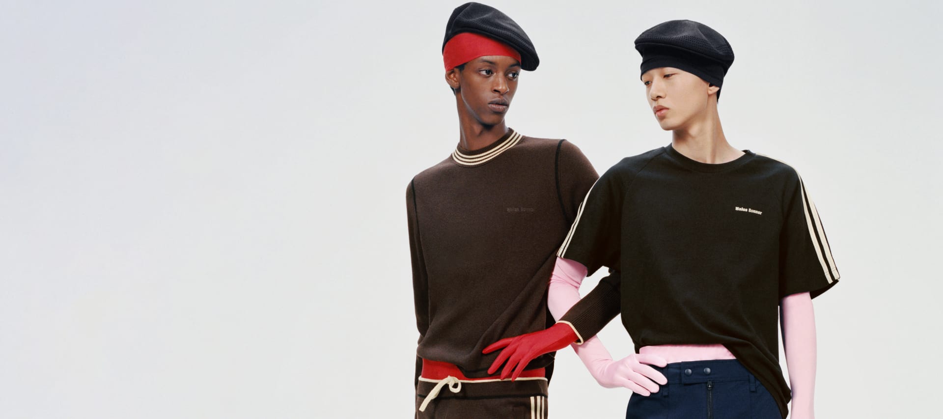 Two models are posing with their arms interlocked against a grey background, wearing apparel from the adidas Originals by Wales Bonner collection.