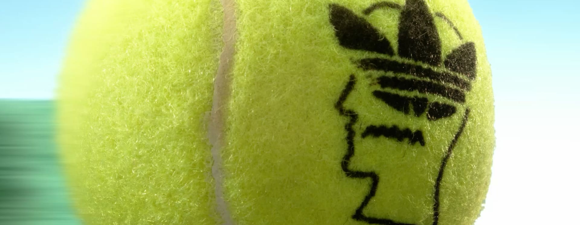 A close-up of a tennis ball stamped with the Brain Dead and adidas logos, set against a blurred green background.