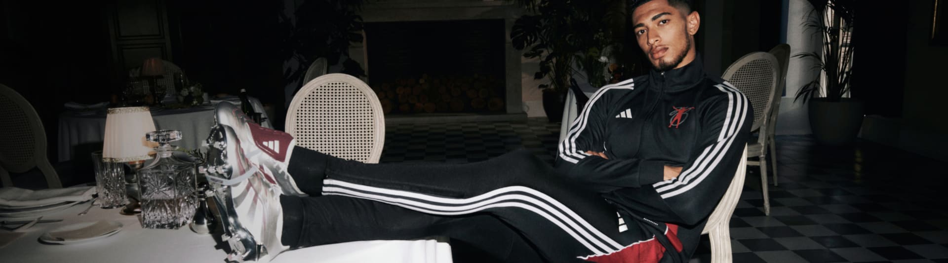 Jude Bellingham wearing a black and white Adidas tracksuit is standing in front of a dark background, with a large white object, possibly an appliance or piece of furniture, visible behind them.
