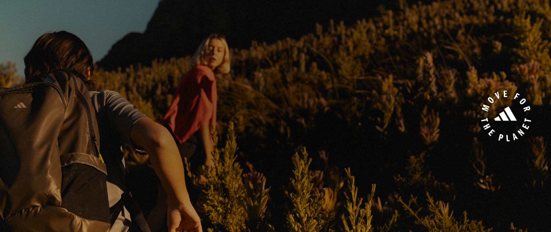 The image shows two people, a woman in a red dress and a man, standing in a field of tall grass against a dark background.
