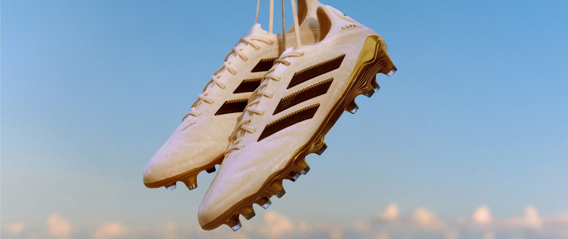 A pair of white and gold COPA soccer cleats suspended against a clear blue sky.