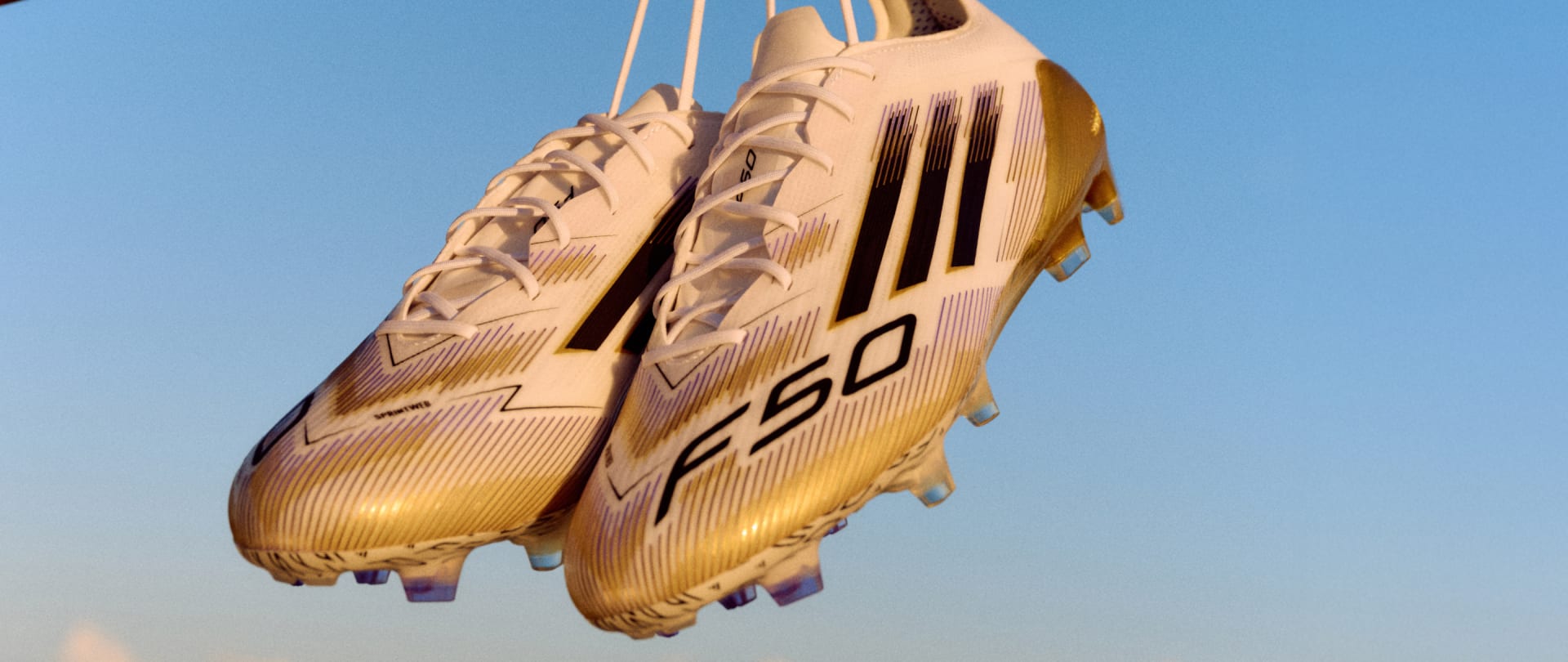 A pair of gold-colored soccer cleats with distinctive patterns and the "F50" logo are suspended against a clear blue sky.