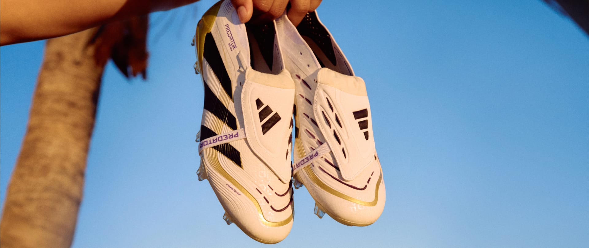 The image shows a pair of white and black soccer boots with distinctive patterns and designs, held up against a clear blue sky background.