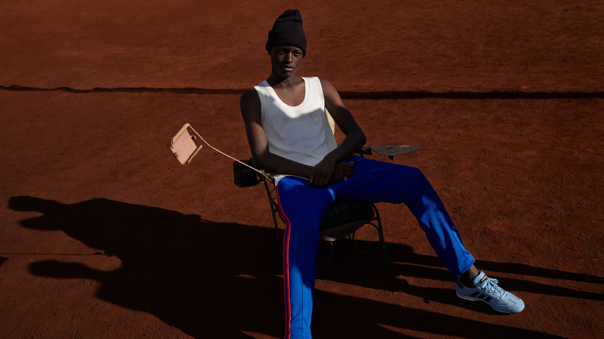 A model poses sitting in a chair in the sun with their legs stretched out, wearing pieces from the adidas Originals by Wales Bonner collection.
