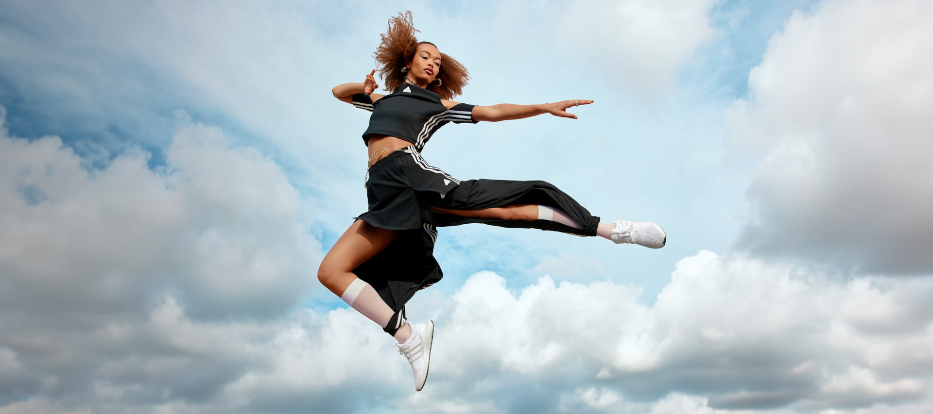 Female model with curly hair jumping into the air. She is wearing a black adidas crop top with cut out shoulders, a black adidas mini skirt over black adidas track pants with a slit in the front, and white socks and sneakers.