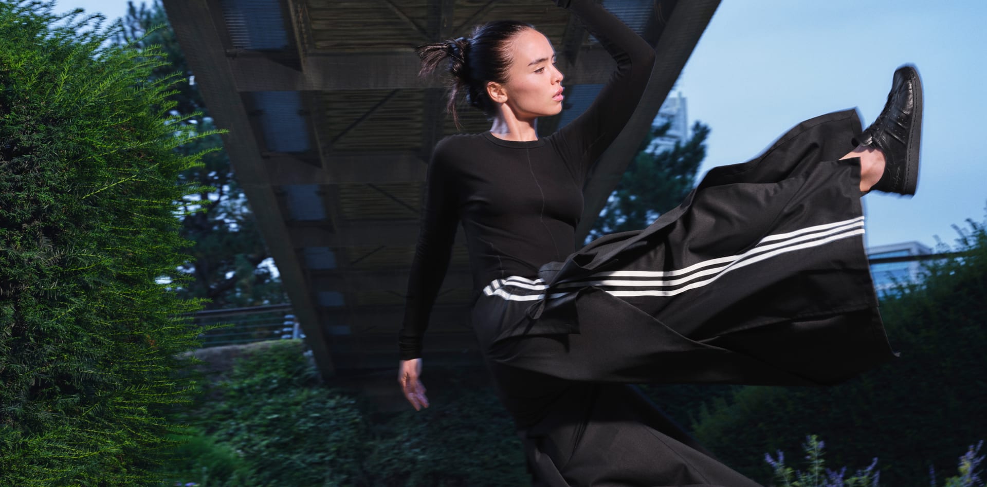 A woman in black Y-3 apparel stands in nature under a concrete bridge, kicking her feet high into the air.