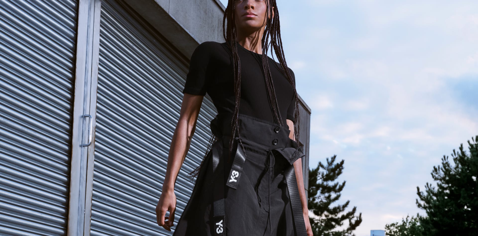A woman with black braids wears all-black Y-3 apparel while posing in the city.