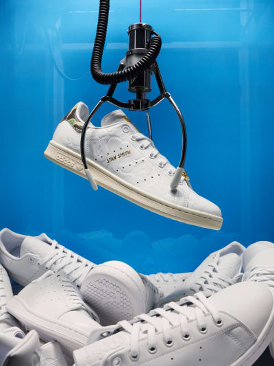 The adidas Originals x BAPE® Stan Smith sneaker is shown hanging from a claw inside an arcade game with a blue backdrop and multiple white Stan Smith's piled below.