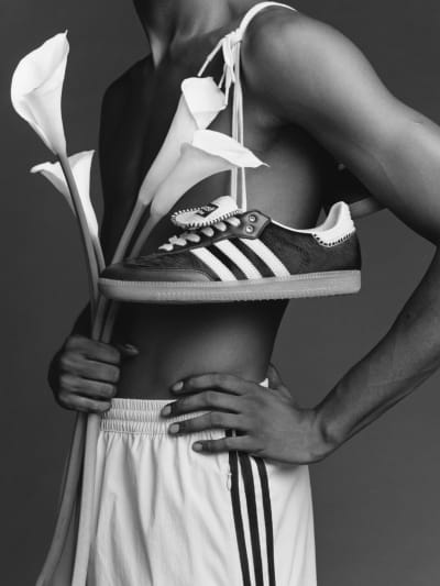 A model is posing wearing white trackpants, with flowers in one hand, and the other hand on their hip. A pair of adidas Originals by Wales Bonner Samba's are hanging over their shoulder.