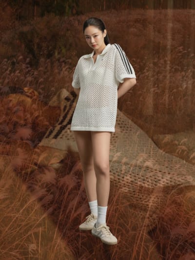 Woman wearing white mesh top, white socks and cream adidas sneakers with white stripes stands, superimposed on photo of an autumnal field.