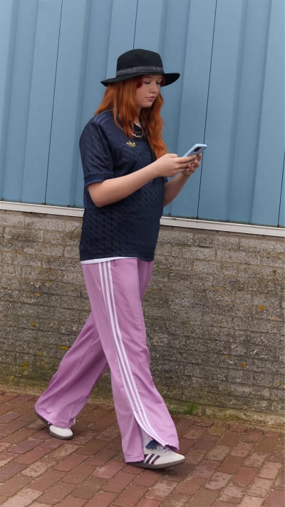 A young woman with long, red hair wearing a black hat and a dark sweater stands on a brick walkway in front of a gray metal wall, holding a mobile device in her hands.