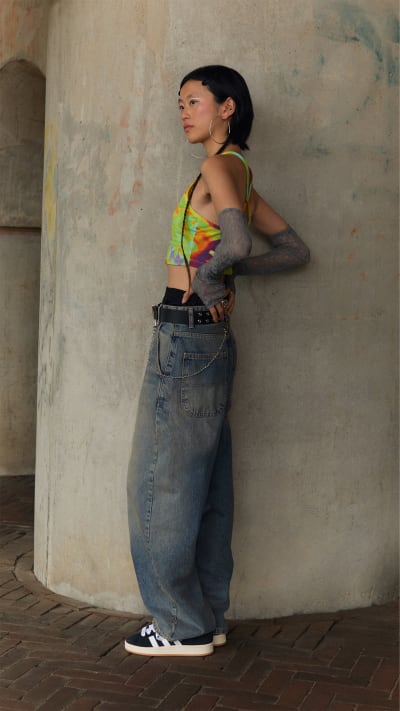 A young person wearing a neon green crop top and baggy jeans stands against a weathered concrete wall.