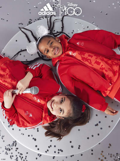 Two children from birds-eye view lying on a circular podium with confetti around them. Both wearing the adidas | Disney100 collection.