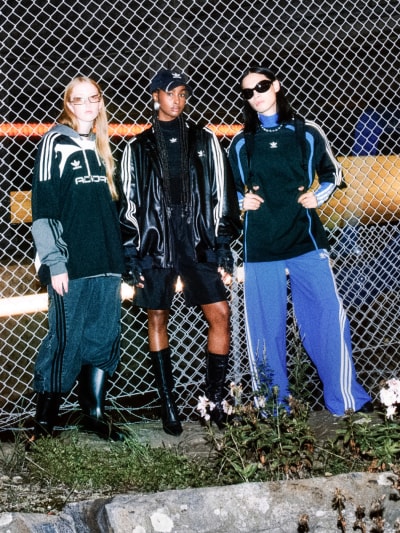 Three people in adidas Adilenium track pants, jackets, and caps lean casually against a steel fence at night while looking into the camera.