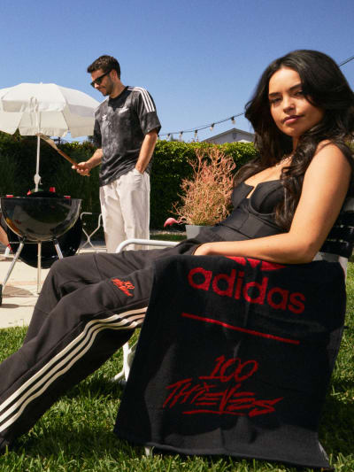 A group of young men and women dressed in adidas Originals x 100 Thieves apparel enjoying a barbecue in a sunny backyard.